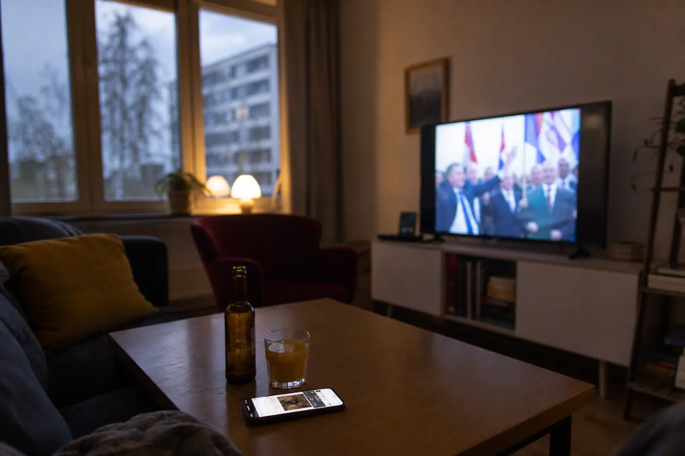 Olohuoneen pöydällä on puhelin, televisio ja kaksi erilaista juomaa; taustalla näkyy epätarkka uutislähetys.