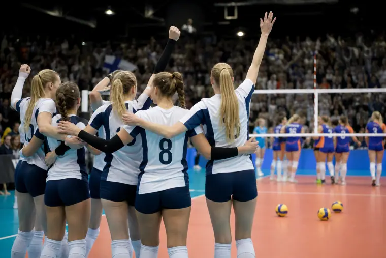 Sinivalkoisiin väreihin pukeutuneita lentopalloilijoita juhlimassa verkon lähellä sisähallissa.